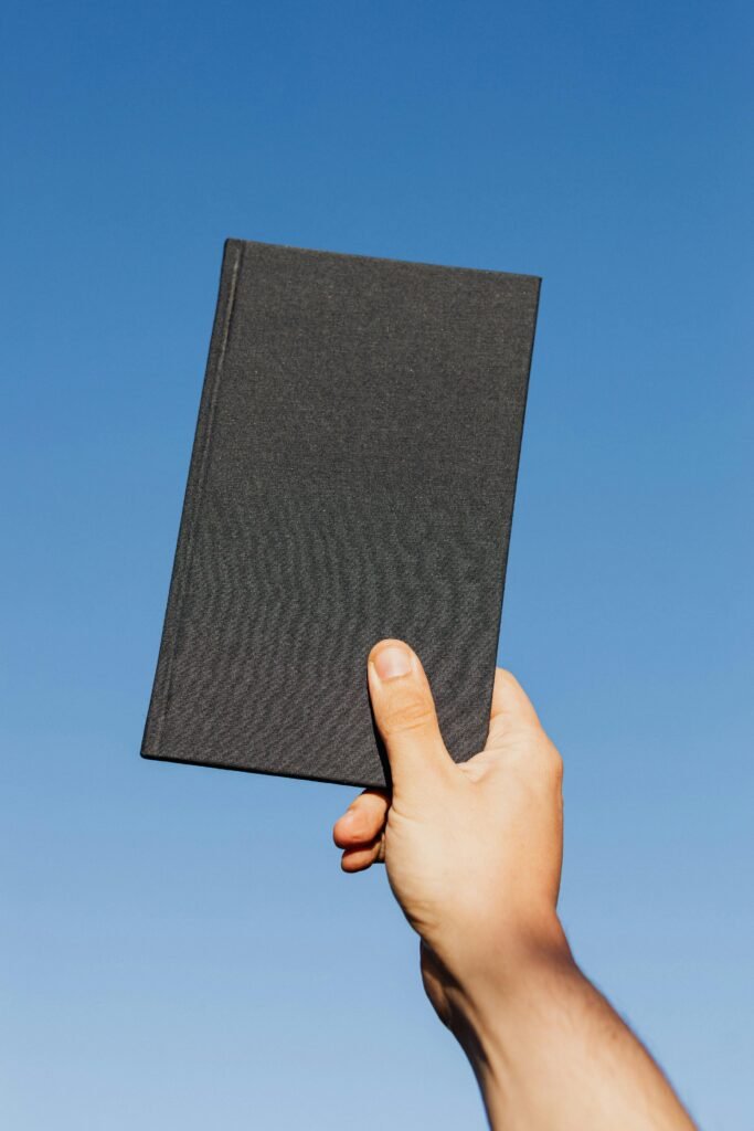 Crop unrecognizable person with closed black notebook in hand against clear blue sky